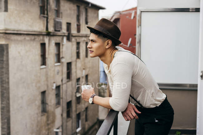 Homem de chapéu bebendo café enquanto se inclina no corrimão da varanda — Fotografia de Stock