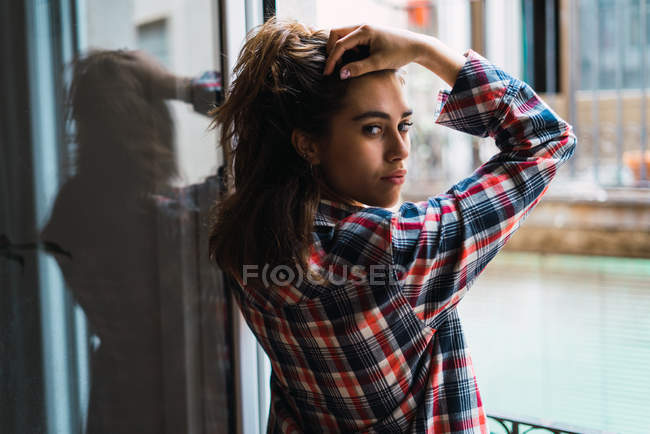 Mulher jovem segurando o cabelo e olhando para trás para a câmera pela janela . — Fotografia de Stock
