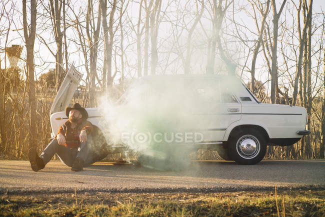 Vista lateral do homem com chapéu apoiado em carro quebrado emitindo fumaça na beira da estrada . — Fotografia de Stock