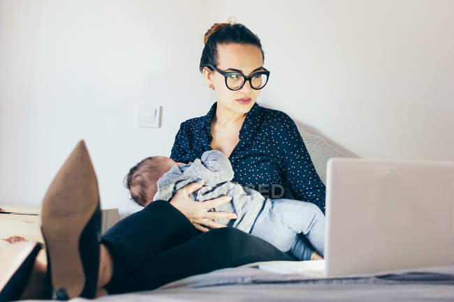 Brunette woman in glasses holding sleeping child and browsing laptop — Stock Photo