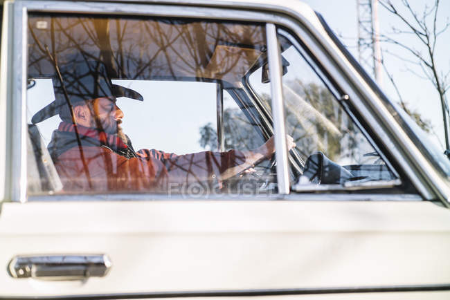Vista lateral do homem barbudo em chapéu dirigindo carro branco na natureza . — Fotografia de Stock