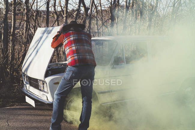 Man in checkered shirt opening hood of smoking broken car in nature. — Stock Photo