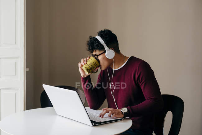 Jovem bebendo café enquanto digita no laptop em casa — Fotografia de Stock