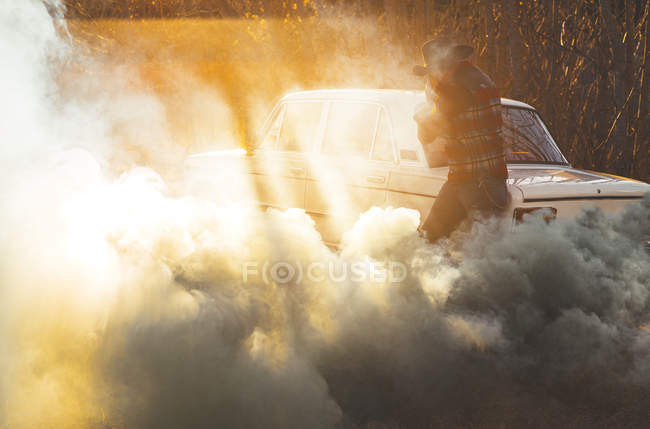 Вид збоку людини з капелюхом ховається обличчям в хмарі диму з розбитого автомобіля під час заходу сонця — стокове фото