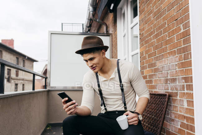 Чоловік з чашкою перегляду смартфона на балконі — стокове фото