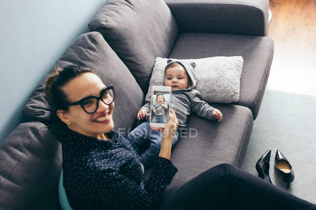 From above cheerful woman looking at camera and showing smartphone with toddler son shot. — Stock Photo