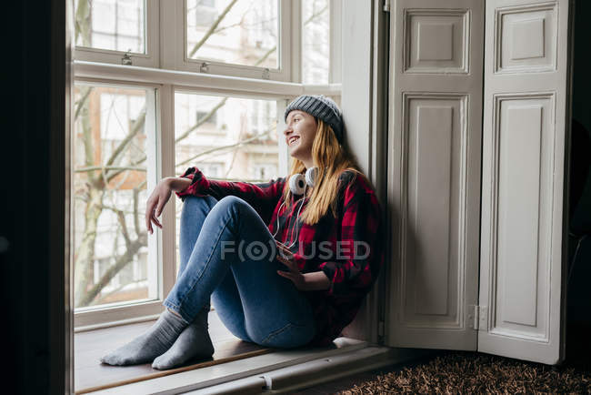Giovane donna sorridente con le cuffie sul collo seduta alla finestra — Foto stock