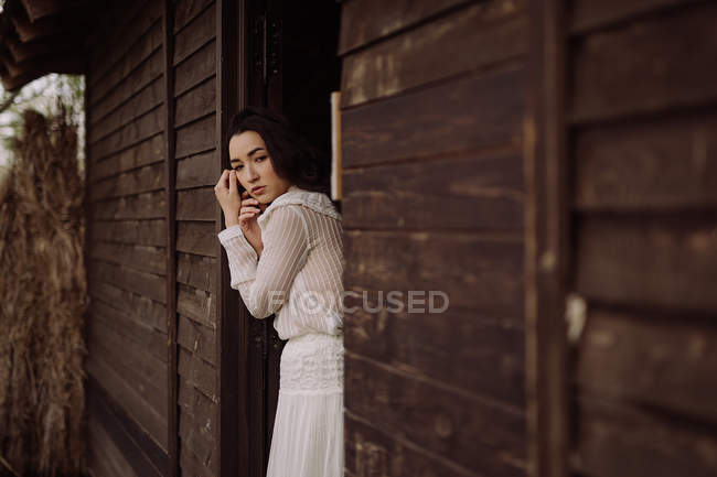 Jovem morena delicada em vestido branco inclinando-se na porta de madeira e olhando para longe . — Fotografia de Stock