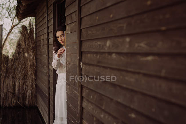 Молода тендітна брюнетка в білій сукні дивиться з дерев'яного дверного отвору — стокове фото