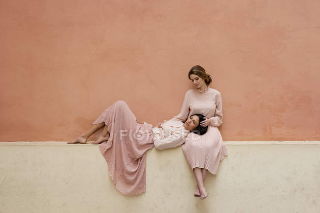 Brunette woman lying on knees of girlfriend sitting at orange wall — Stock Photo