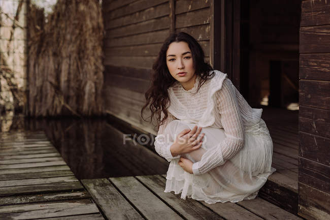 Young brunette in white dress sitting on doorstep of wooden cabin — Stock Photo