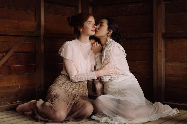 Young brunettes wearing old-fashioned clothes and sitting in tender embrace on wood. — Stock Photo