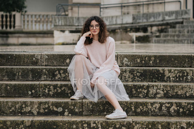 Brunette in glasses and veil skirt sitting on wet stairs and looking at camera — Stock Photo