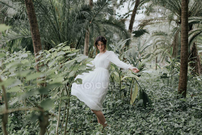 Brunette woman in white light dress spinning around among green tropical plants. — Stock Photo