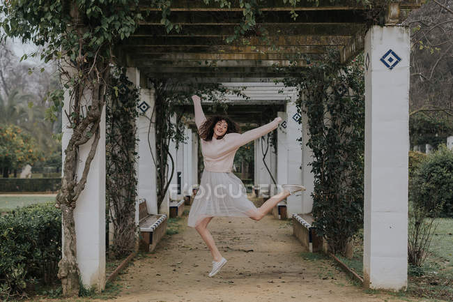 Захоплена дівчина стрибає в алеї з зеленими повзаючими рослинами на білих колонах . — стокове фото