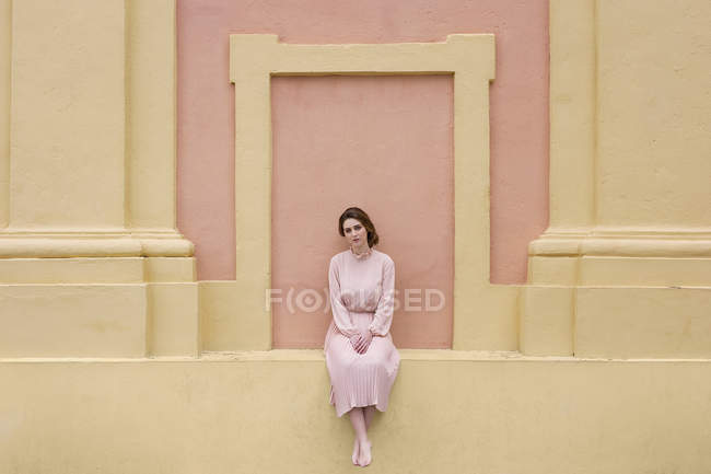 Женщина в розовом платье сидит у розовой стены и смотрит в камеру — стоковое фото