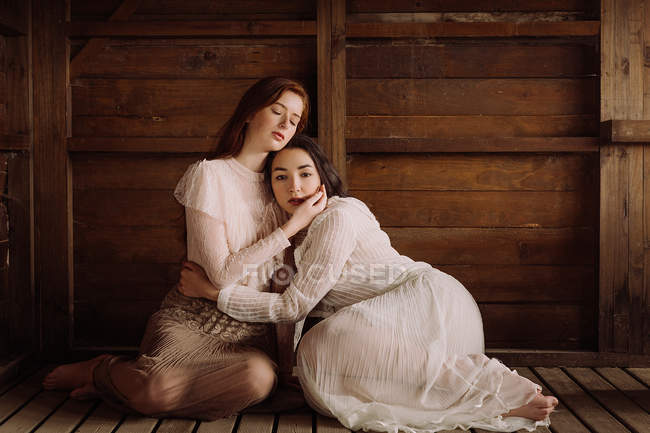 Young brunettes wearing old-fashioned elegant clothes and posing in tender embrace on wood. — Stock Photo
