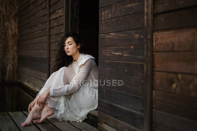 Jeune brune en robe blanche assise à la porte en bois avec les yeux fermés — Photo de stock