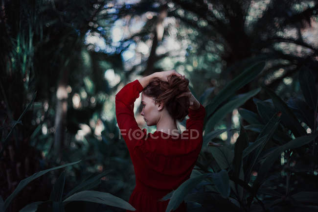 Visão traseira da menina segurando o cabelo no pão e olhando para longe entre o verde exuberante . — Fotografia de Stock