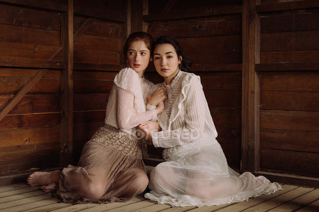 Young brunettes wearing old-fashioned elegant clothes and embracing at wooden cabin — Stock Photo