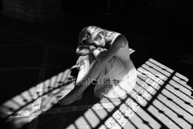 Mujer en vestido blanco claro sentado en el suelo de baldosas a la sombra de las persianas y mirando a la cámara . - foto de stock