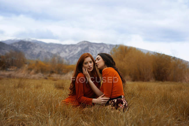 Young girls in affection sitting on dry grass with mountains on background. — Stock Photo