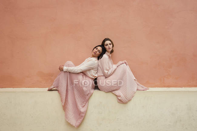 Two women in vintage dresses sitting back to back at orange wall. — Stock Photo