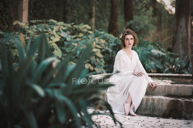 Young brunette woman in white clothes sitting at fountain in park. — Stock Photo