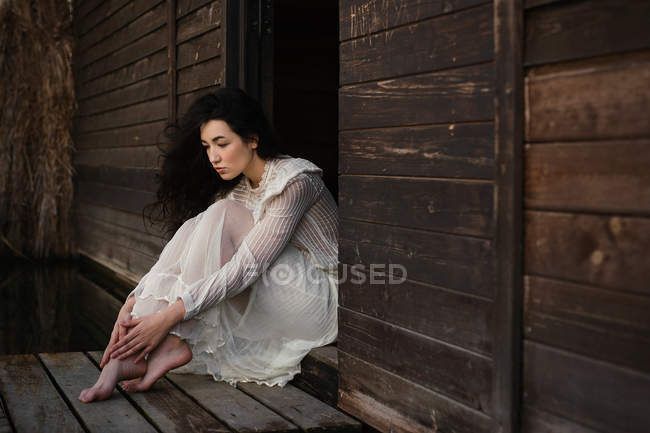 Молодая нежная брюнетка в белом платье сидит у деревянных дверей — стоковое фото