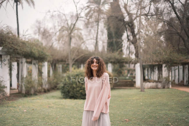 Усміхнена кучерява брюнетка в светрі і спідниці позує на газоні в садовій алеї . — стокове фото