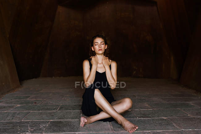 Junge Frau im schwarzen Kleid sitzt barfuß und mit geschlossenen Augen im steinernen Raum. — Stockfoto