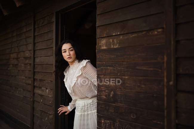 Joven morena vestida de blanco asomada a la puerta de madera - foto de stock