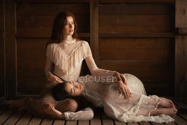 Young girls wearing old-fashioned elegant clothes and posingin tender embrace on wood. — Stock Photo
