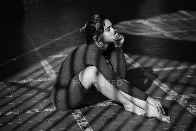 Sensual chica sosteniendo la barbilla en la mano y sentado en el suelo en los rayos del sol - foto de stock