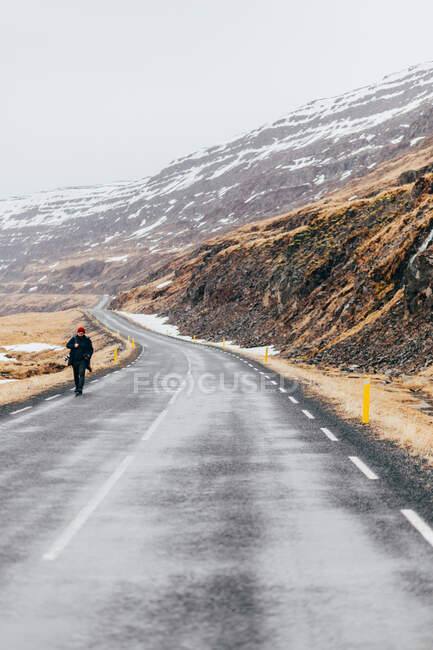 Persona con zaino che cammina da sola su una lunga carreggiata sotto montagne rocciose nella fredda Islanda. — Foto stock