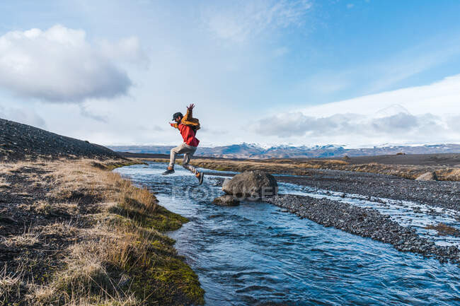 Бічний погляд на юнака у випадковому одязі, який стрибає через струмок, подорожуючи чудовою ісландською сільською місцевістю. — стокове фото