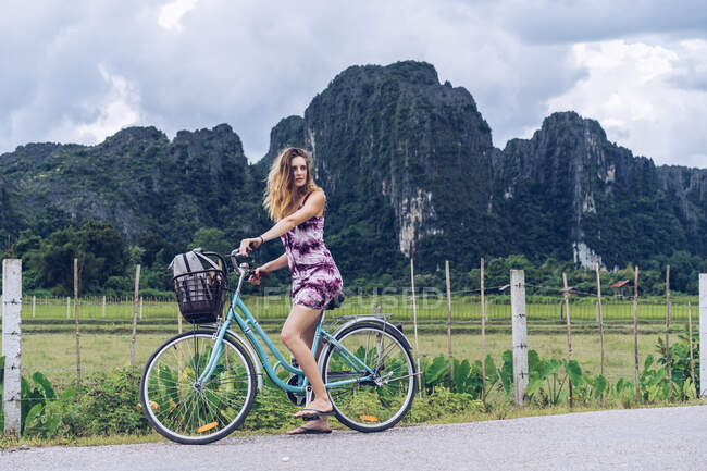 Pretty young woman riding on bicycle on rural road on background of mountains. — Stock Photo