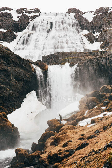 Vista a distanza dell'uomo in piedi sul bordo roccioso della collina con cascata schizzi sullo sfondo, Islanda. — Foto stock