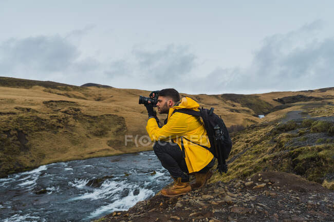 Vue latérale du jeune photographe utilisant un appareil photo pour faire des photos de la magnifique campagne islandaise tout en étant assis près du ruisseau — Photo de stock