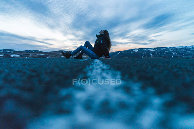 Vista lateral jovem mulher em roupas quentes sentado no meio da estrada de asfalto enquanto viaja pela Islândia. — Fotografia de Stock