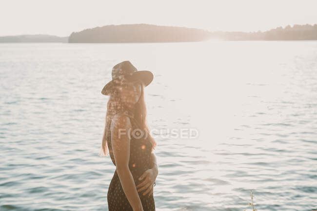 Seitenansicht einer lässigen Frau mit Hut, die im grellen Sonnenlicht gegen gleißendes Wasser steht und in die Kamera blickt — Stockfoto