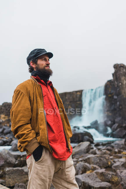 Hombre barbudo guapo mirando hacia otro lado mientras está de pie en el fondo de la hermosa cascada durante el viaje a través de Islandia. - foto de stock