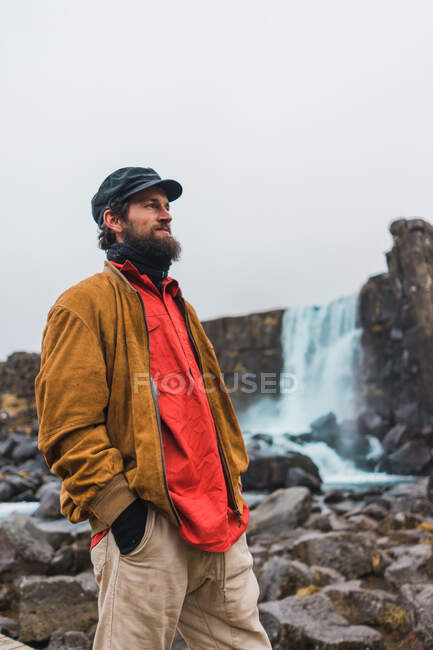 Bello uomo barbuto guardando lontano mentre in piedi sullo sfondo di una bella cascata durante il viaggio attraverso l'Islanda. — Foto stock