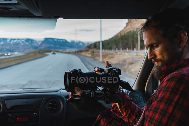 Bärtiger Mann im karierten Hemd sitzt auf dem Beifahrersitz des Autos und macht Fotos während einer Reise durch Island. — Stockfoto