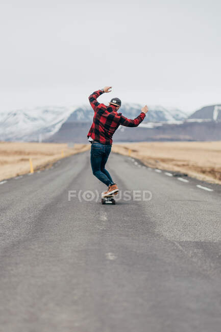 Vista trasera del chico hipster montando monopatín en camino largo pavimentado con montañas nevadas en el fondo en Islandia. - foto de stock