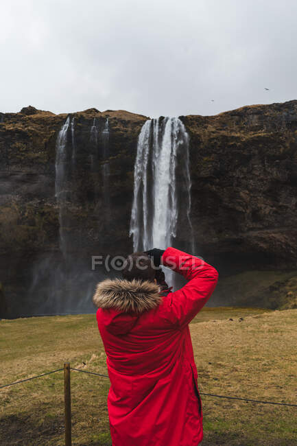 Rückansicht eines Mannes in warmer Kleidung mit professioneller Kamera, um schöne Wasserfälle in Island zu fotografieren — Stockfoto