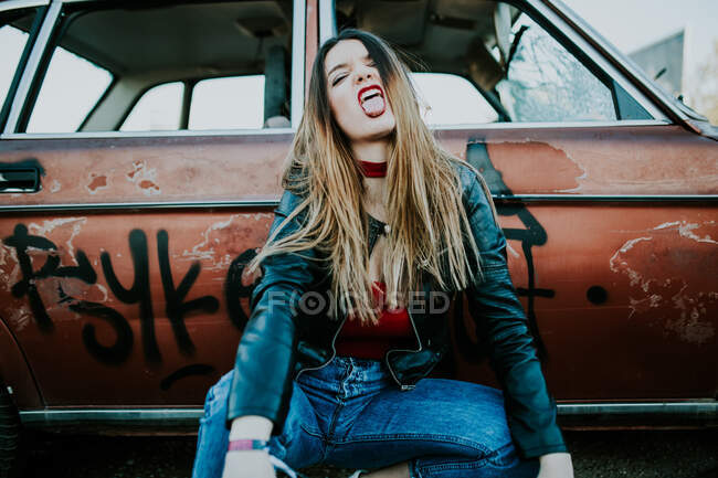Attractive glamour woman looking at camera and showing tongue out while sitting at grungy vintage car. — Stock Photo