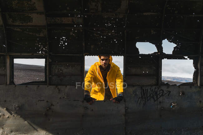 Joven guapo con chaqueta amarilla parado fuera de la carcasa del avión abandonado y mirando a la cámara mientras viaja a través de Islandia - foto de stock