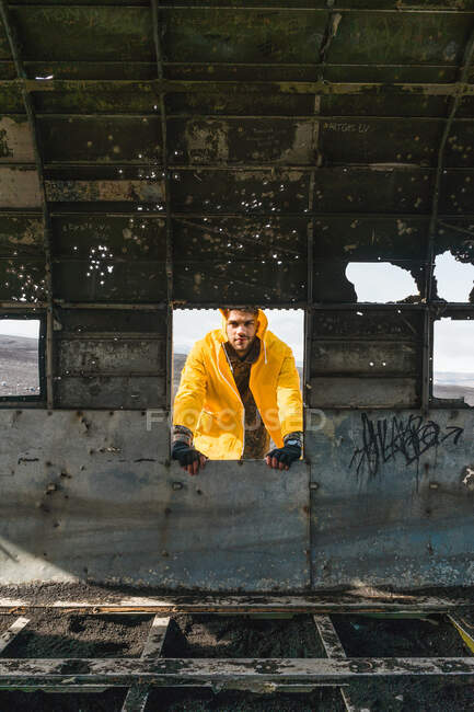 Пристрасний молодий чоловік у жовтій куртці стоїть біля покинутого тулуба літака і дивиться на камеру під час подорожі по Ісландії. — стокове фото