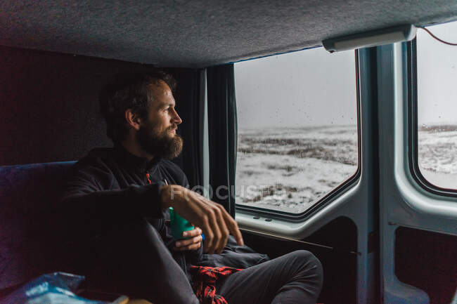 Handsome bearded man holding cup of hot drink and looking out window while traveling in comfortable van through magnificent Iceland. — Stock Photo